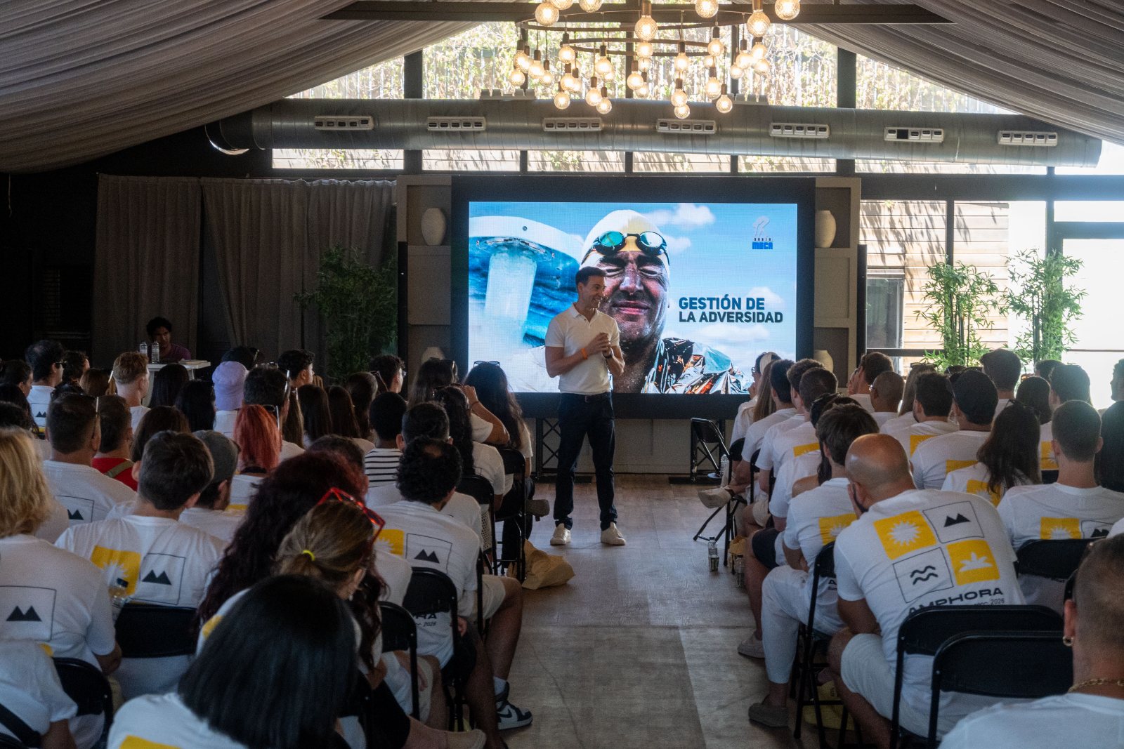 Keynote speaker addressing a seated audience in branded t-shirts at the Amphora Summer Meet
