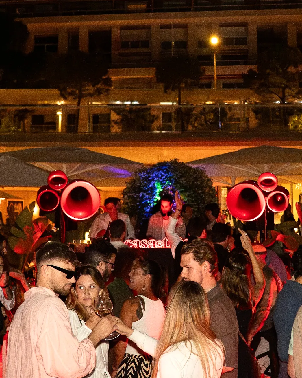 Night at Neptune Beach Club: a packed crowd in front of the DJ booth, red horn speakers and palms lit with fairy lights